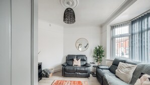 A modern living room with a black leather sofa, a black leather armchair, a round mirror, and a large window with curtains. - Sleeps7-pet&familyfriendly With Parking (Coventry)