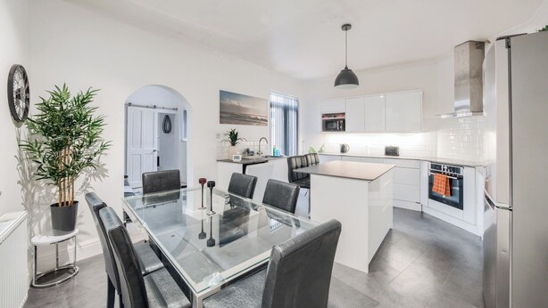 A modern kitchen with white cabinets, a central island, and a dining area with a glass table and chairs. - Sleeps7-pet&familyfriendly With Parking (Coventry)