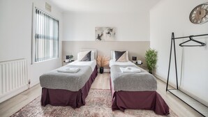 A modern bedroom with two beds, a large window, a radiator, a clock, and a hanging rack. - Sleeps7-pet&familyfriendly With Parking (Coventry)