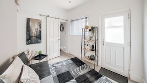 A modern living room with a black and white rug, a sofa with cushions, a black metal shelf with decorative items, and a cityscape painting on the wall. - Sleeps7-pet&familyfriendly With Parking (Coventry)