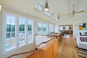 A spacious living area with a wooden staircase, large windows, a dining area, and a kitchen.