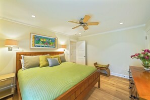 Una camera da letto con un letto, una ventola a soffitto, un comodino, una lampada, una porta e un quadro appeso al muro.