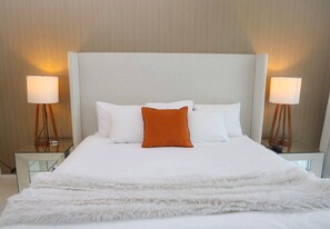 Ein Hotelzimmer mit einem großen Bett, weißer Bettwäsche, einem orangefarbenen Zierkissen und zwei Nachttischlampen.