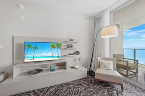 A hotel room with a large TV on a white stand, a floor lamp, a sofa, a view of the ocean, and a balcony with a chair.