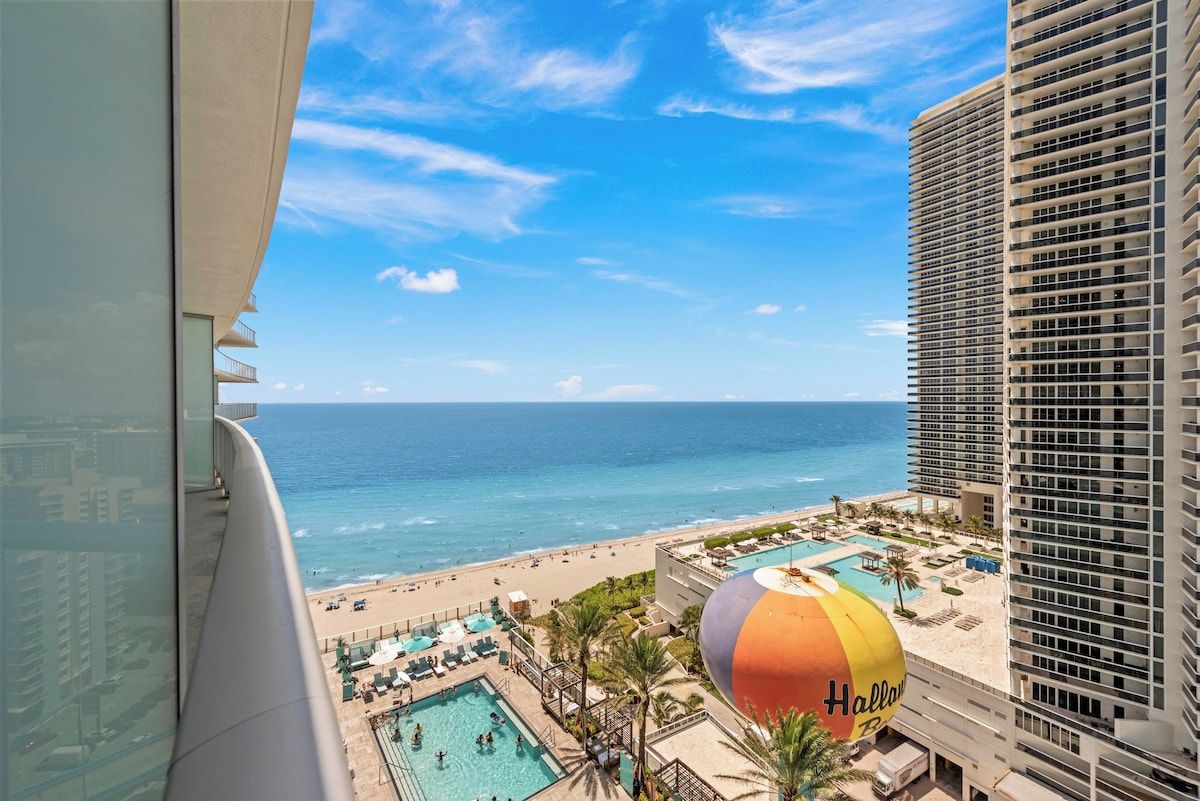 Een hotelbalkon met uitzicht op een strand, een zwembad en een luchtballon.