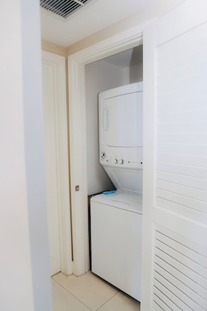 A laundry room with a front-loading washing machine and dryer, a door leading to another room, and a tiled floor.