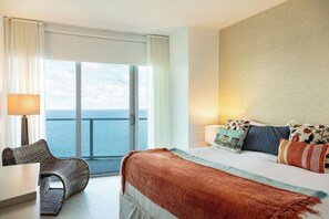 A hotel room with a bed, a chair, a desk, and a view of the sea.