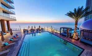 A rooftop pool with lounge chairs and a palm tree, overlooking the ocean.
