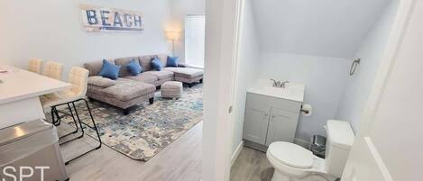 Sala de estar con un sofá, sillas y alfombra. También se muestra un baño con inodoro, lavamanos y armarios.