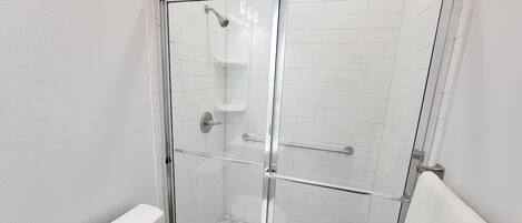 Ducha con cerramiento de vidrio, pared de azulejos blancos, puerta de cristal y una manija de metal.
