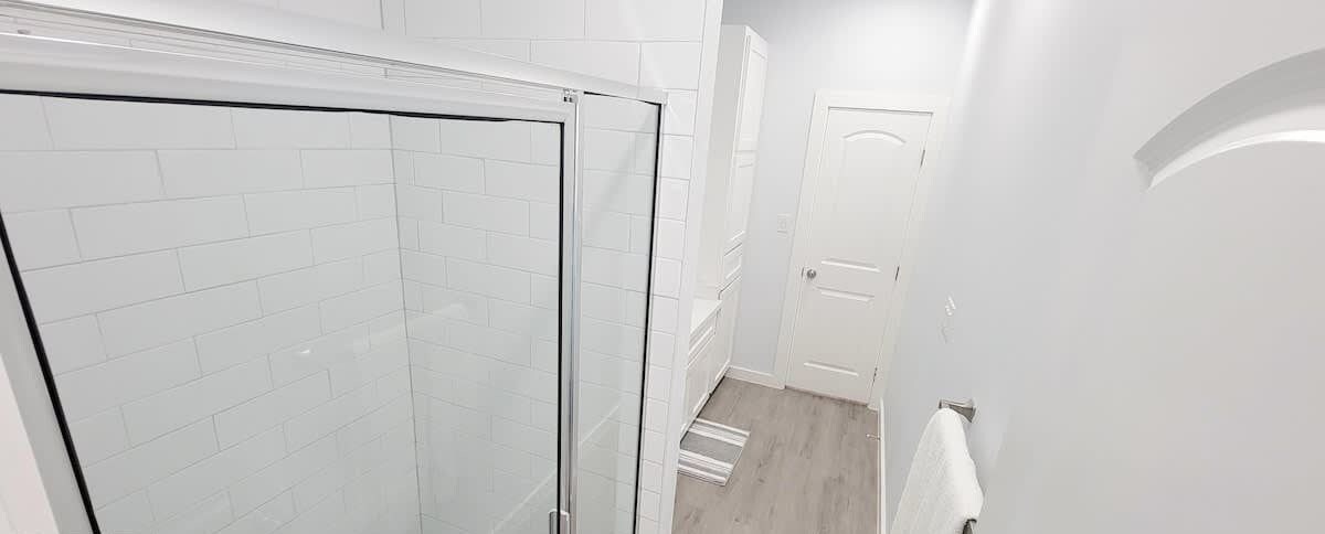 A bathroom with white tiled walls, a glass shower enclosure, and a wooden floor.