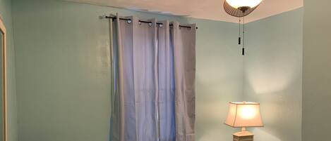 A bedroom with a bed, a nightstand, a lamp, a ceiling fan, and a window with curtains.