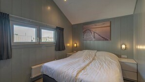 Un dormitorio con una cama grande, dos mesitas de noche, una ventana con cortinas, un radiador y una lámpara fijada a la pared.