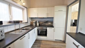 A compact kitchen with white cabinets, a black countertop, a built-in oven, and a window with curtains. - Ferienhaus im Skandinavischen Stil (Rechlin)