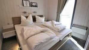 A bedroom with two beds, white bedding, a wooden headboard, a small bedside table, a shelf with two framed pictures, and a window with curtains. - Ferienhaus im Skandinavischen Stil (Rechlin)