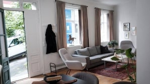 A modern living room with a grey sofa, a white armchair, and a coffee table. There are two windows with curtains, a door leading outside, and a small plant on the coffee table. - In the Heart of Brosundet! Fully Equipped 3 Bed Room Ap (Ålesund)