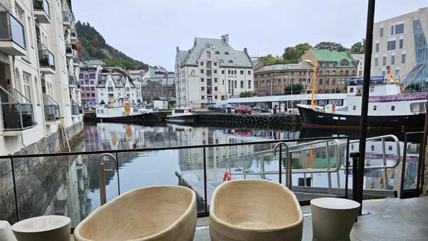 A waterfront terrace with large, beige, bowl-shaped seating and a small table, overlooking a marina with boats and historic buildings. - In the Heart of Brosundet! Fully Equipped 3 Bed Room Ap (Ålesund)