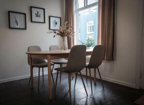 Uma área de refeições com mesa de madeira e quatro cadeiras, quadros botânicos nas paredes e vista de um edifício pela janela.