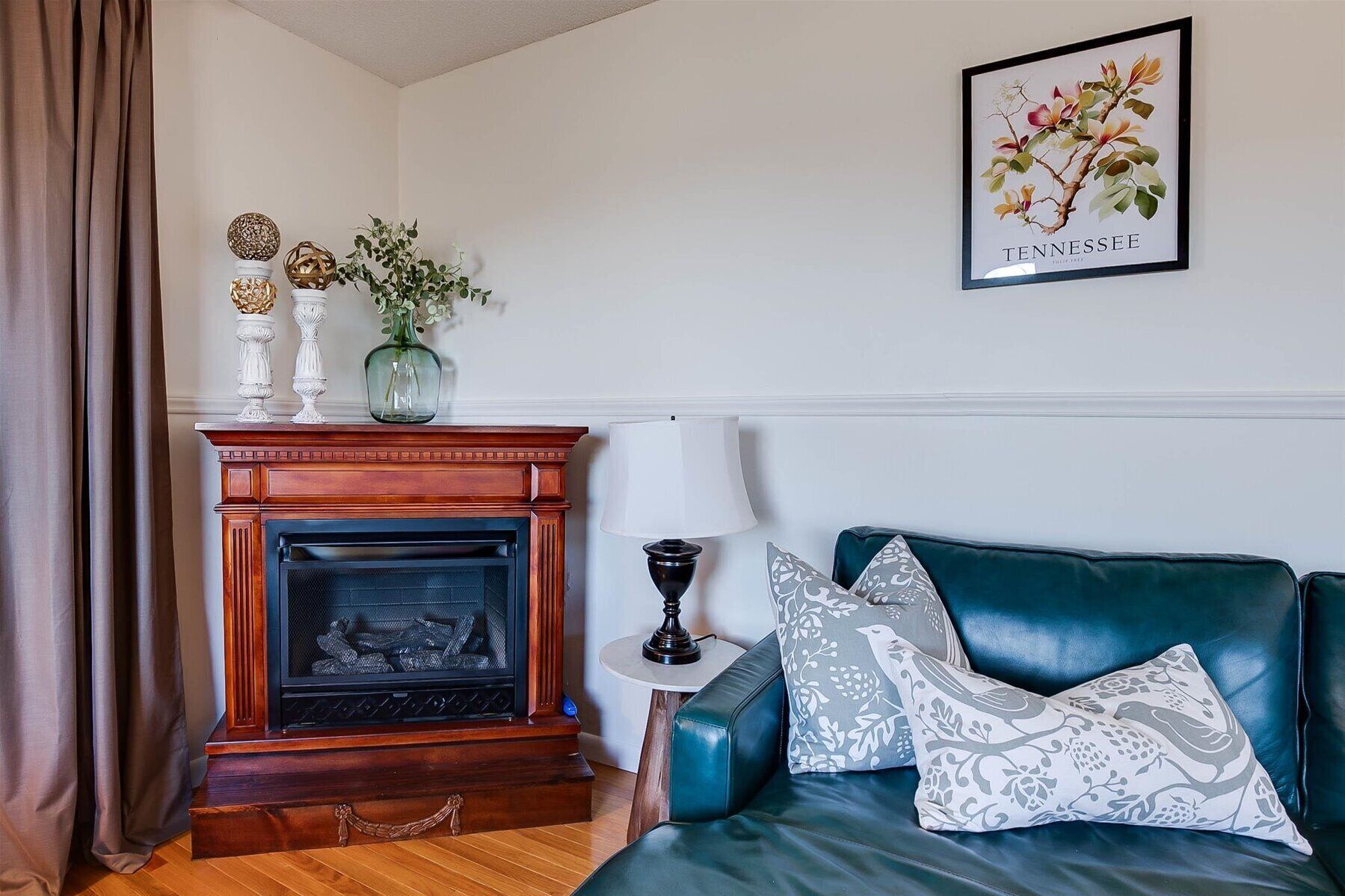 A living room with a fireplace, a green sofa with patterned cushions, and a framed botanical print on the wall.