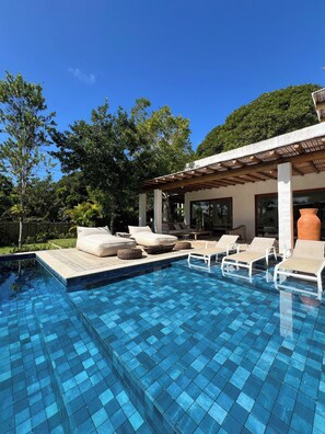 A swimming pool with tiled blue floor, surrounded by lounge chairs and cushions.