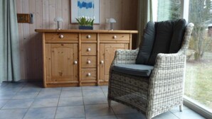 A wicker chair with a cushion, a wooden cabinet with drawers, a framed abstract painting, and a window with a view of trees. - Ferienhaus mit Sauna im Ferienpark Mirow (Mirow)