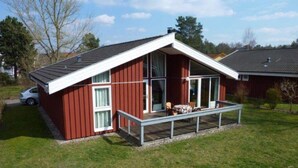 A red building with a sloped roof, a covered porch with chairs, and a car parked nearby. - Ferienhaus mit Sauna im Ferienpark Mirow (Mirow)