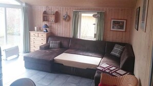 Una sala de estar acogedora con un sofá seccional, una mesita, paredes de madera, una ventana con cortinas y un ventilador de techo.
