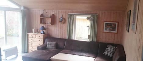A cozy living room with a sectional sofa, a small table, wooden walls, a window with curtains, and a ceiling fan.