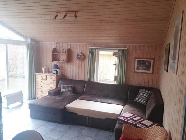 Una sala de estar acogedora con un sofá seccional, una mesita, paredes de madera, una ventana con cortinas y un ventilador de techo.