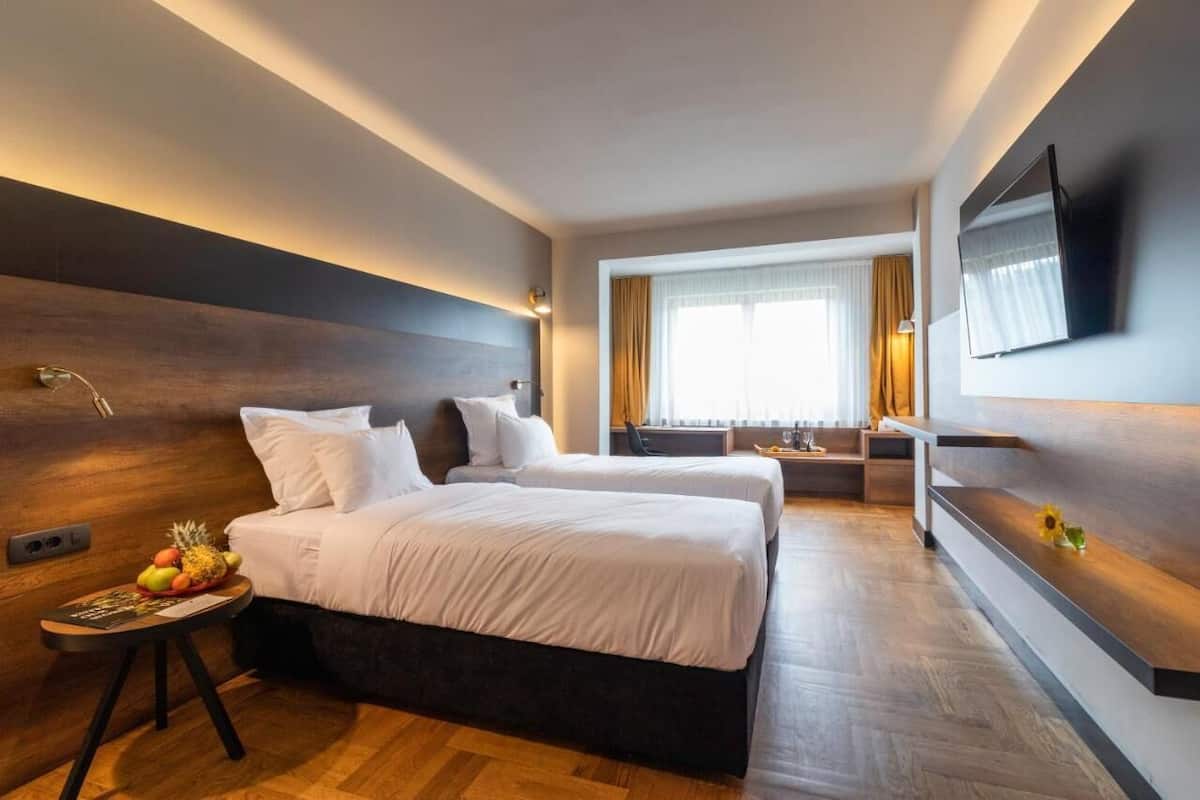 Una habitación de hotel moderna con dos camas, un televisor de pantalla plana, un cabecero de madera, una mesita con frutas y una ventana con cortinas.