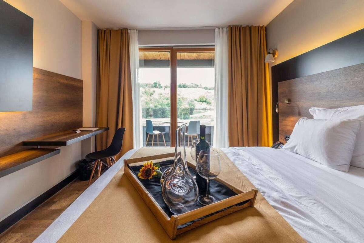 Habitación de hotel con cama, mesita de noche, bandeja con botella de vino y copa, silla y vistas al exterior.