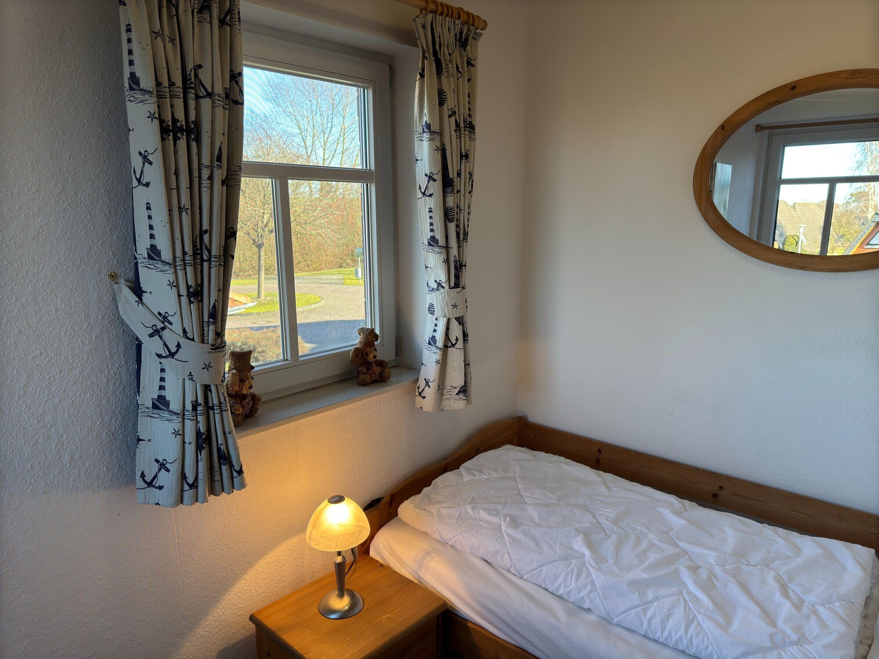 A bedroom with a bed, a nightstand with a lamp, a window with curtains, and a mirror on the wall.