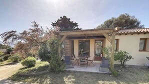 Un espace extérieur aménagé, abrité et doté d’une table et de chaises, entouré d’un jardin où poussent différentes plantes et un petit arbre.