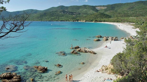 Une plage côtière aux eaux turquoise et limpides, bordée de rochers et d’une étendue de sable.