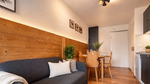 En moderne stue med en mørkegrå sofa, træpaneler på væggen, et spiseområde med et træbord og stole, og et køkken med en opvaskemaskine.