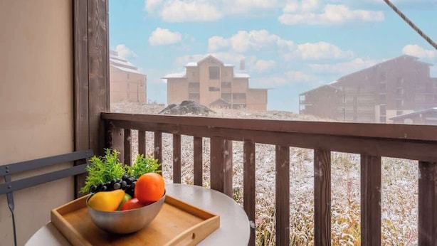 En balkon med gelænder af træ, et bord med en skål frugt og udsigt til bygninger i det fjerne.