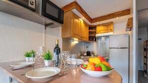 A kitchen with wooden cabinets, a microwave, and a refrigerator. There is a dining table with plates, cutlery, and a bowl of fruit.