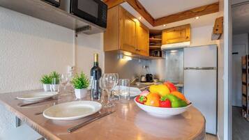 Een keuken met houten kasten, een magnetron en een koelkast. Er is een eettafel met borden, bestek en een schaal fruit.