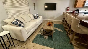 A living room with a sofa, a coffee table, and a TV. There is a kitchen area with a microwave and a coffee machine. - Beachfront Bliss – Steps to Dining, Shops & More! (Biloxi)