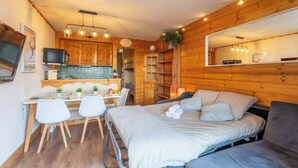 A cozy room with a bed, a desk with chairs, a kitchen area, and a large mirror.