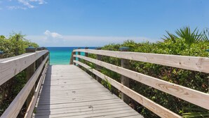 Ein hölzerner Steg führt zu einem Strand mit klarem, blauen Wasser und blauem Himmel.