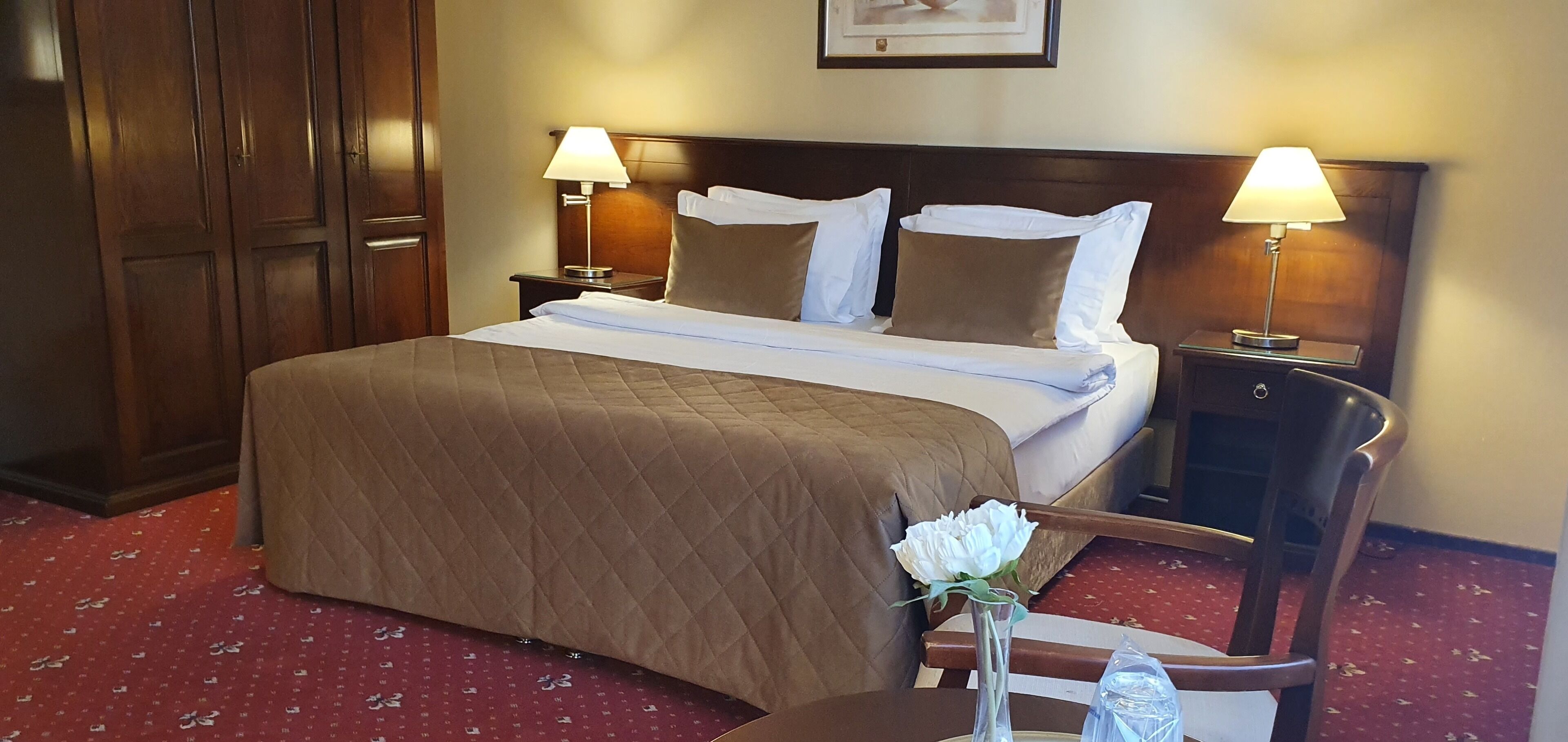 A hotel room with a queen-sized bed, two bedside tables with lamps, a wooden wardrobe, a red carpet, and a vase with a white flower on a small table.