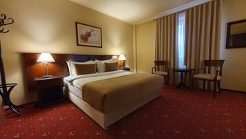Habitación de hotel con una cama grande, dos mesitas de noche con lámparas, una silla, una pequeña mesa y una ventana con cortinas.