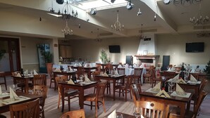 Ein geräumiges Restaurant mit mehreren Tischen und Stühlen, Deckenleuchten und einem im Hintergrund sichtbaren Küchenbereich.