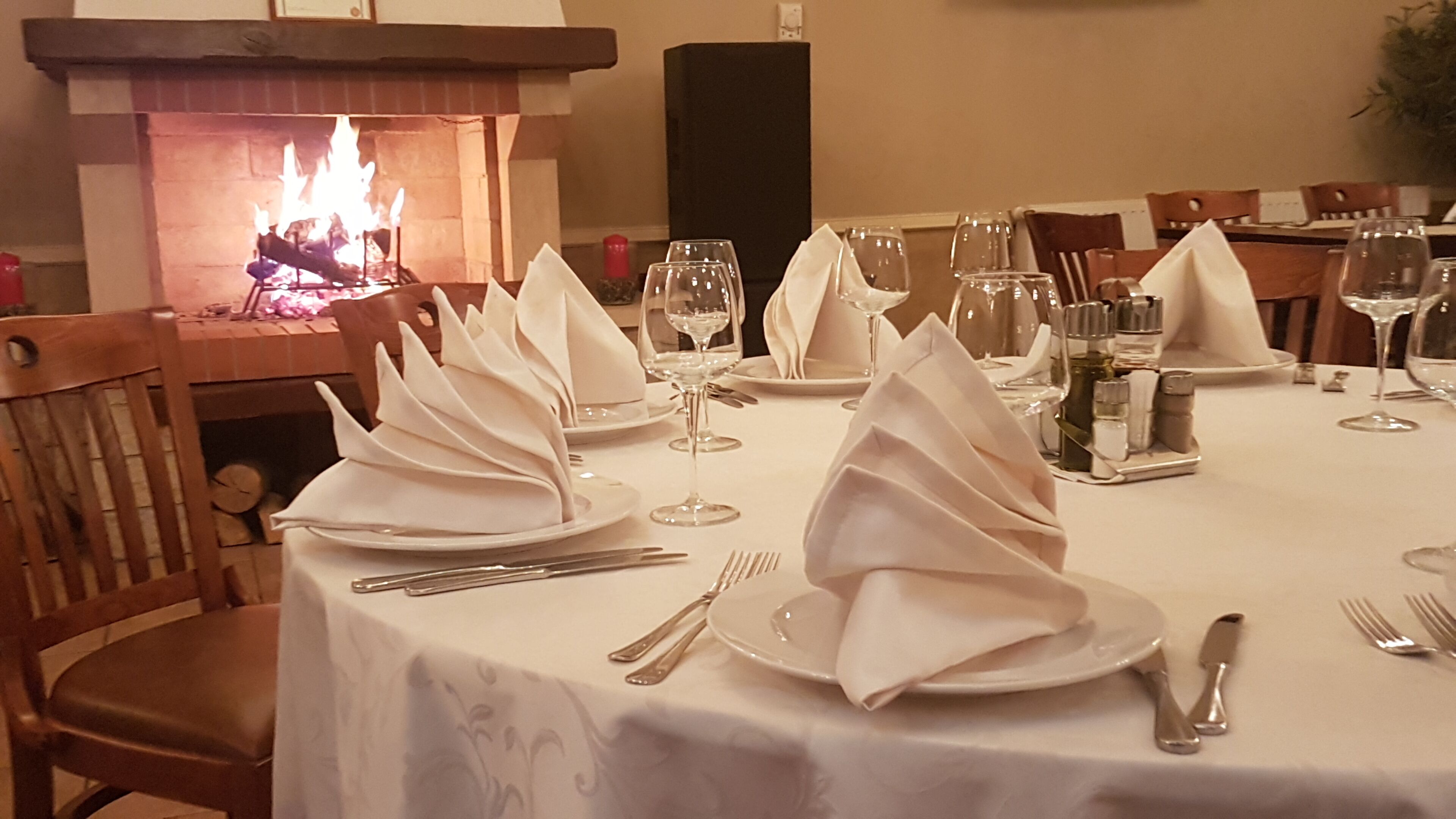 A dining table set for a meal with white napkins, wine glasses, and cutlery. A fireplace is visible in the background.