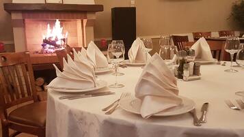 Una mesa de comedor dispuesta para una comida, con servilletas blancas, copas de vino y cubiertos. Se ve una chimenea al fondo.