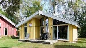 Un edificio amarillo con un amplio ventanal y ventanas, un patio con mobiliario exterior y un edificio rojo al fondo.