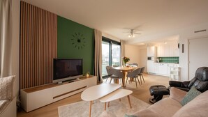 A modern living room with a flat-screen TV, a dining area with a table and chairs, a kitchen with white cabinets, and a large window. - Apartment in De Panne Near the Beach (Koksijde)