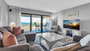 A modern living room with a grey sofa, a white coffee table, and a flat-screen TV. There is a balcony with a view of the sea and a dining area with chairs. - Santa Rosa Dunes 825 (Pensacola Beach)