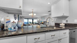 A modern kitchen with white cabinets, a granite countertop, and a stainless steel sink. There are decorative items on the counter and a view of the living area in the background. - Santa Rosa Dunes 825 (Pensacola Beach)